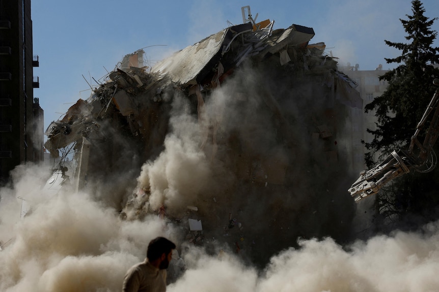 A dark cloud of smoke and dust surrounds a building that is being knocked down. 