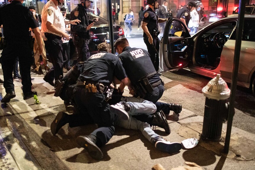 Three uniformed NYPD officers pin a person to the pavement after having removed them from a car, which is nearby.