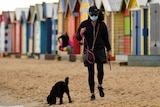 A woman walks her dog past colourful beach huts, wearing a mask, on a grey day