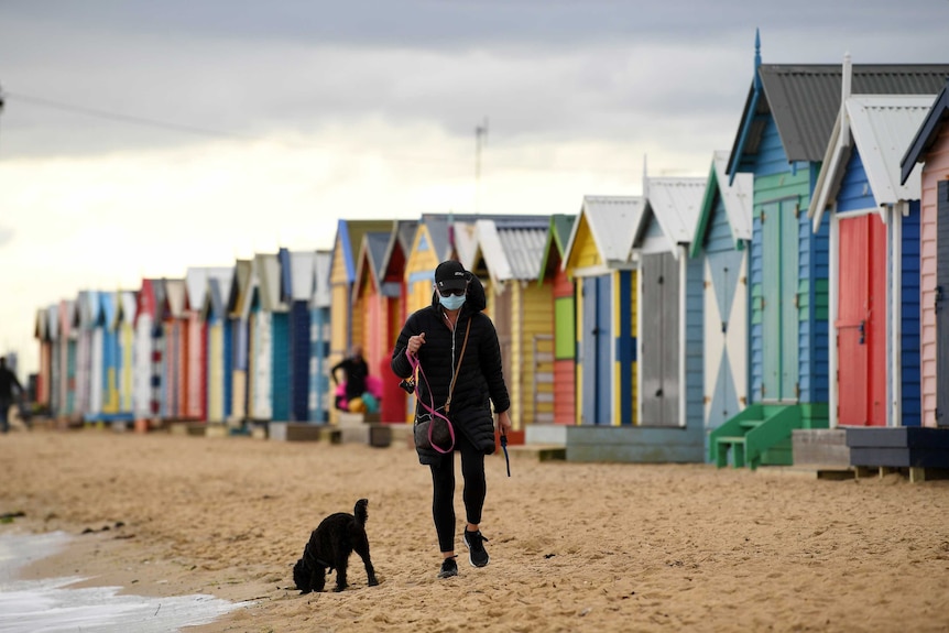 A woman walks her dog past colourful beach huts, wearing a mask, on a grey day