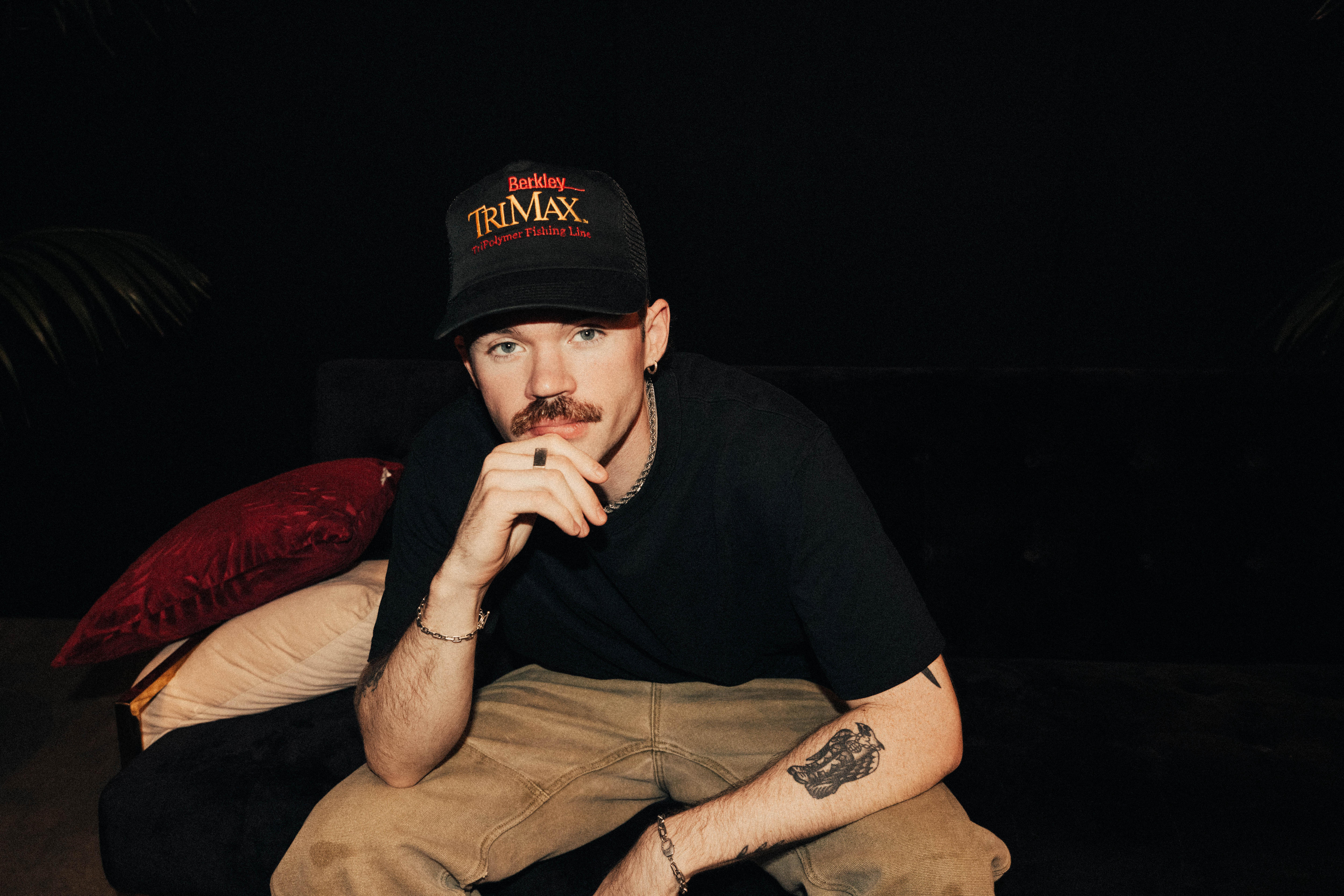 Portrait photo of Dom Dolla sitting in a dark room with a black t shirt, grey jeans and a black trucker hat on
