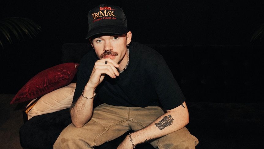 Portrait photo of Dom Dolla sitting in a dark room with a black t shirt, grey jeans and a black trucker hat on