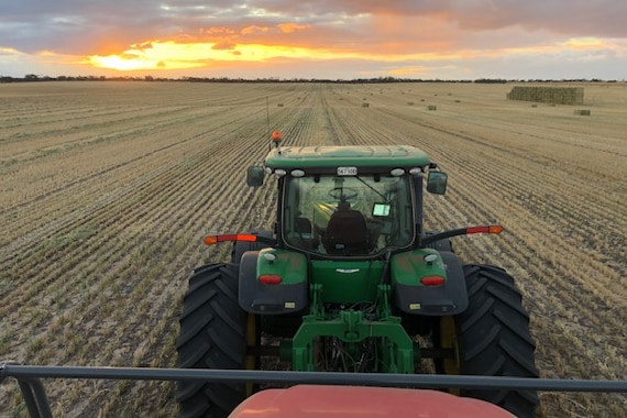 A green tractor is in a freshly harvested field, golden stubble is left behind. Bales sit nearby and the sun sets orange and blu