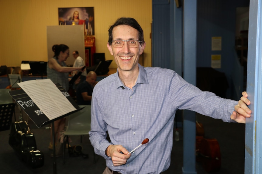 an orchestra conductor leans against a doorframe with a baton smiling at the camera