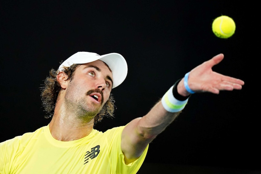 A male tennis player, wearing a cap backwards, holds a tennis ball in the air as he prepares to serve at the Australian Open.