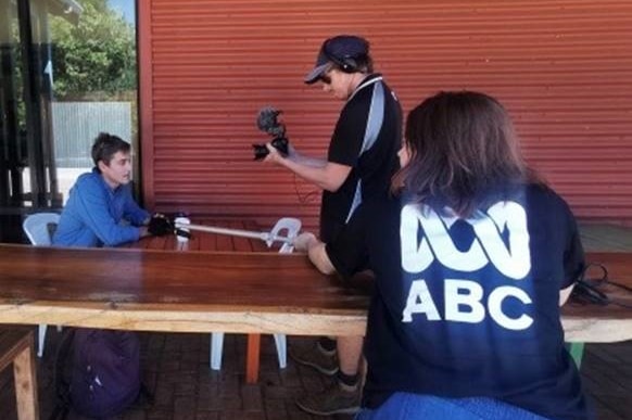 Reporter with ABC logo on back leaning on table holding microphone as cameraman standing filming with small camera.