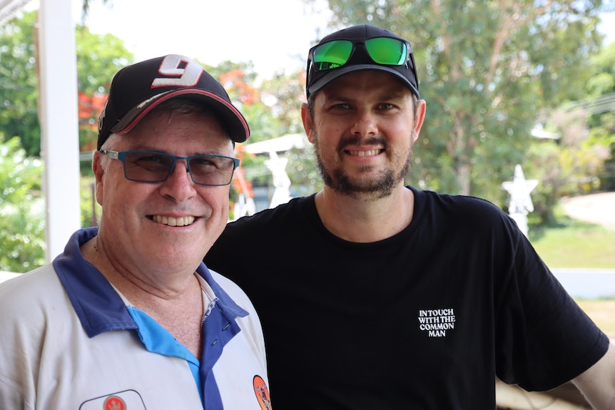 A smiling older man, wearing glasses and a cap, stands next to a younger man, his son.