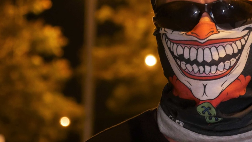A man wearing a bandana with a leering clown's face on it stands on a street at night.