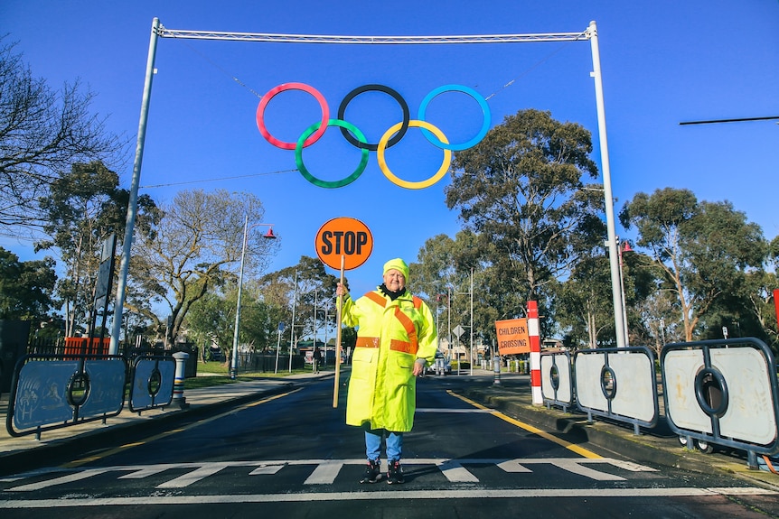 A woman holding a stop sign stands on a pedestrian crossing under Olympic rings.