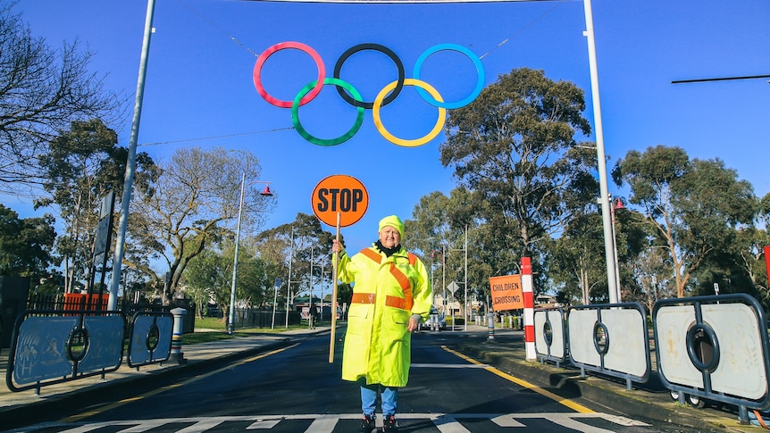 A woman holding a stop sign stands on a pedestrian crossing under Olympic rings.