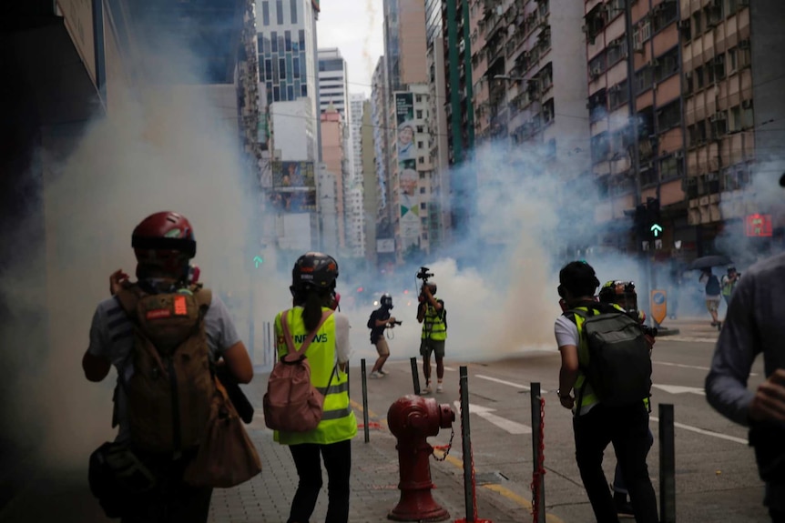 clouds of tear gas flow through a street where media with cameras and masks are standing