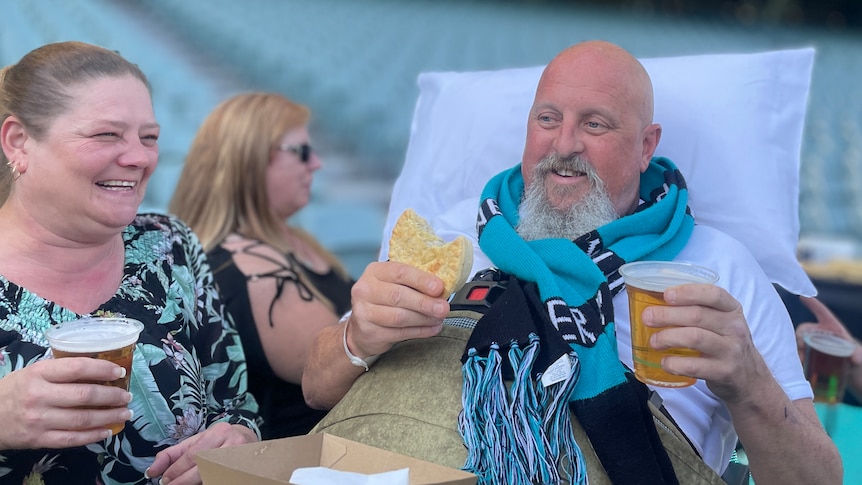 A man in a Port Adelaide footy scarf holding a pie and a beer lying on a stretcher next to two women