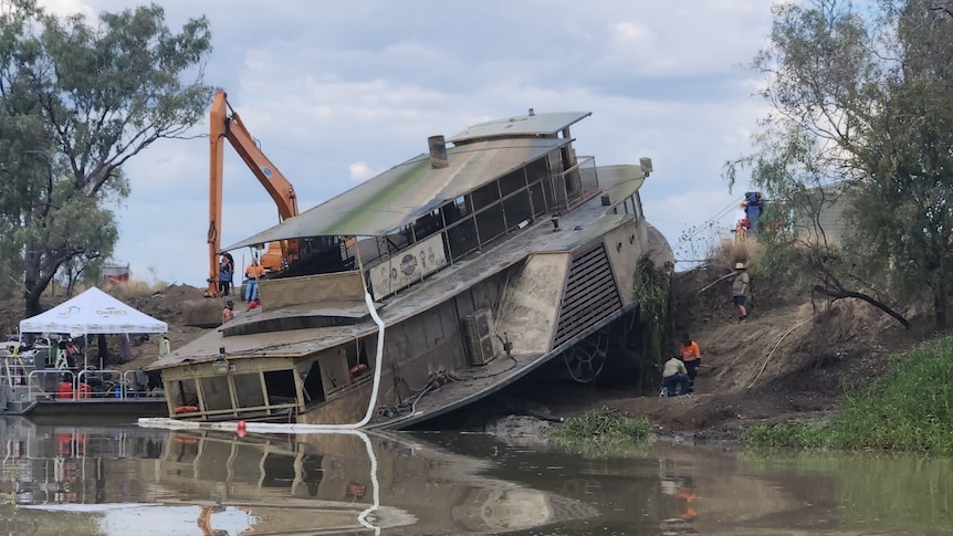 A boat being lifted from a river using cranes, resting half on the the banks.