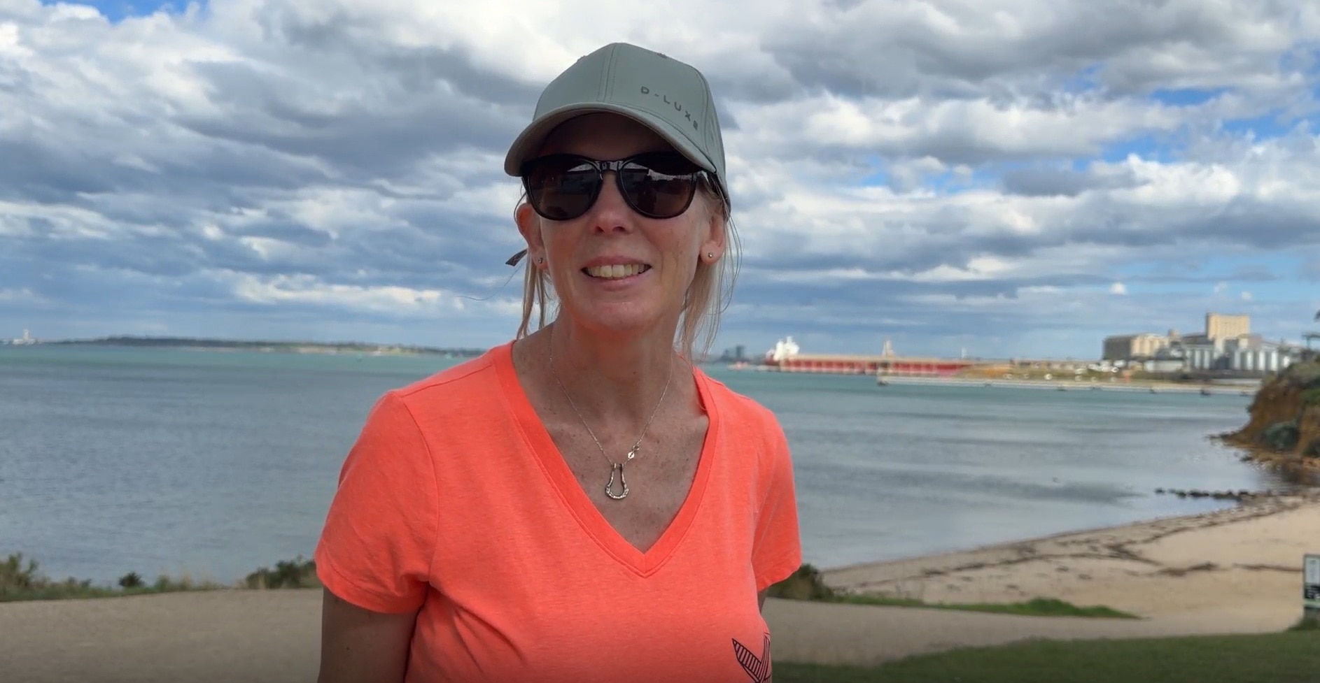 A woman with sunglasses and an orange T-shirt stands on a beach.