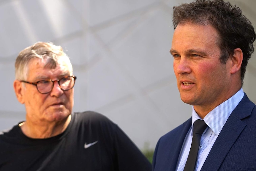 A man in a blue suit and an older man in a grey t-shirt talk outside court.