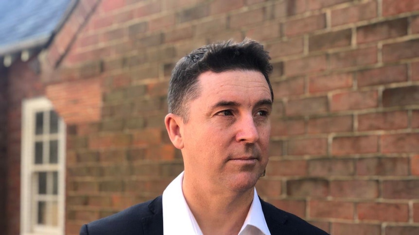 A middle-aged white man in a white shirt and suit jacket looks into the middle distance.