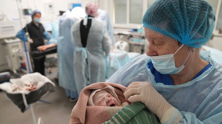 A medical worker in hospital surgery uniform holds and looks down at a newborn wrapped in blankets.