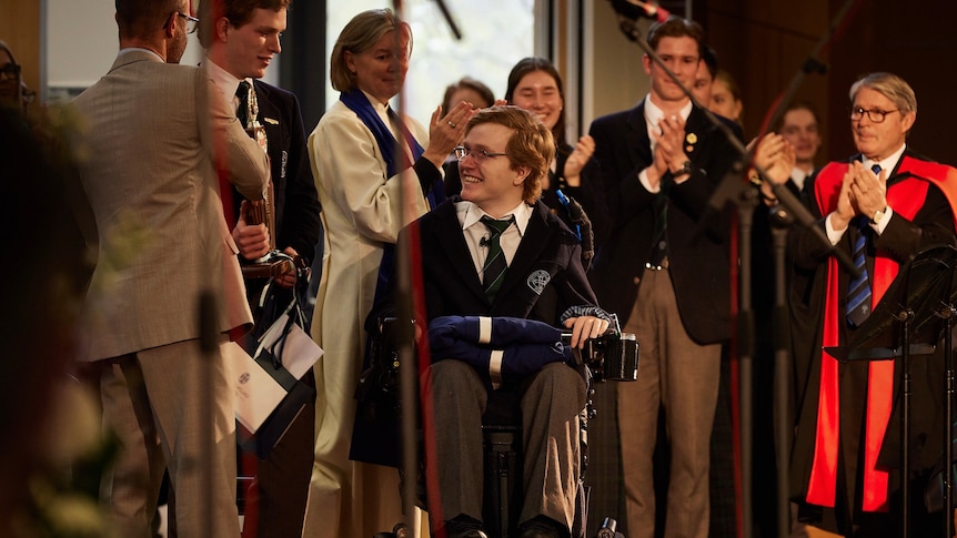 A young man in a wheelchair with other people standing around him clapping.