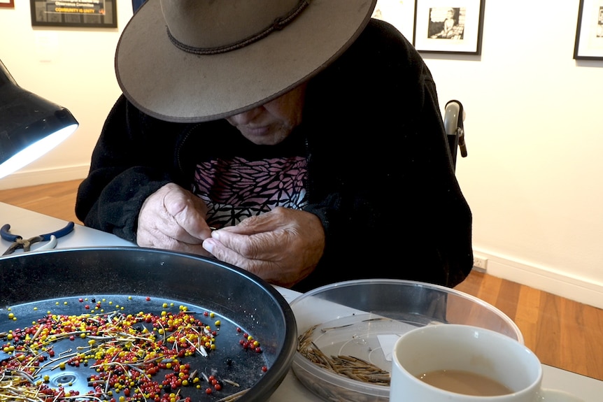 An old man wearing a hat is bent over a tray of beads and echidna spikes