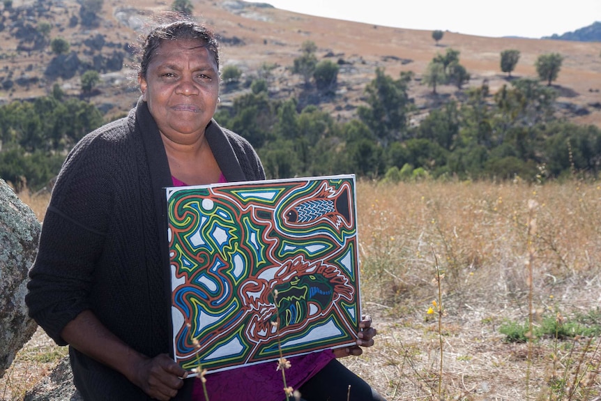 An Aboriginal woman holds a traditional style painting with the background of hills and paddocks