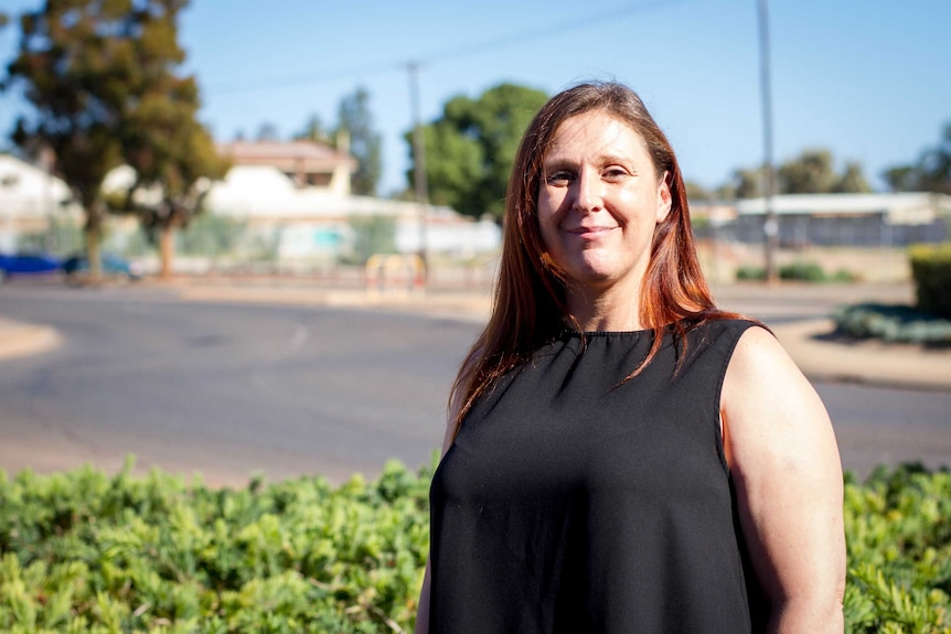 A woman with shoulder-length auburn hair and a black sleeveless top stands in front of a quiet street.