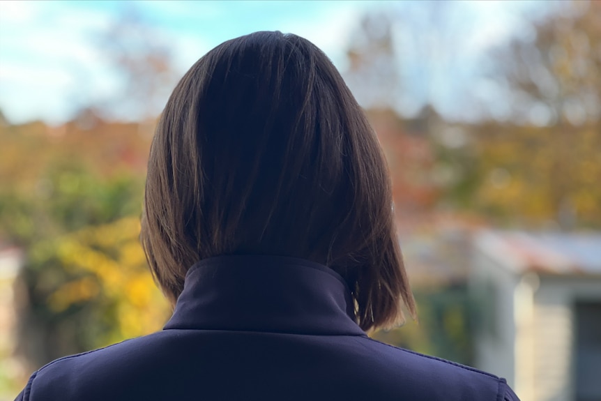 The back of a womans head with dark hair, she is outside the background is blurred but shows trees and a blue sky