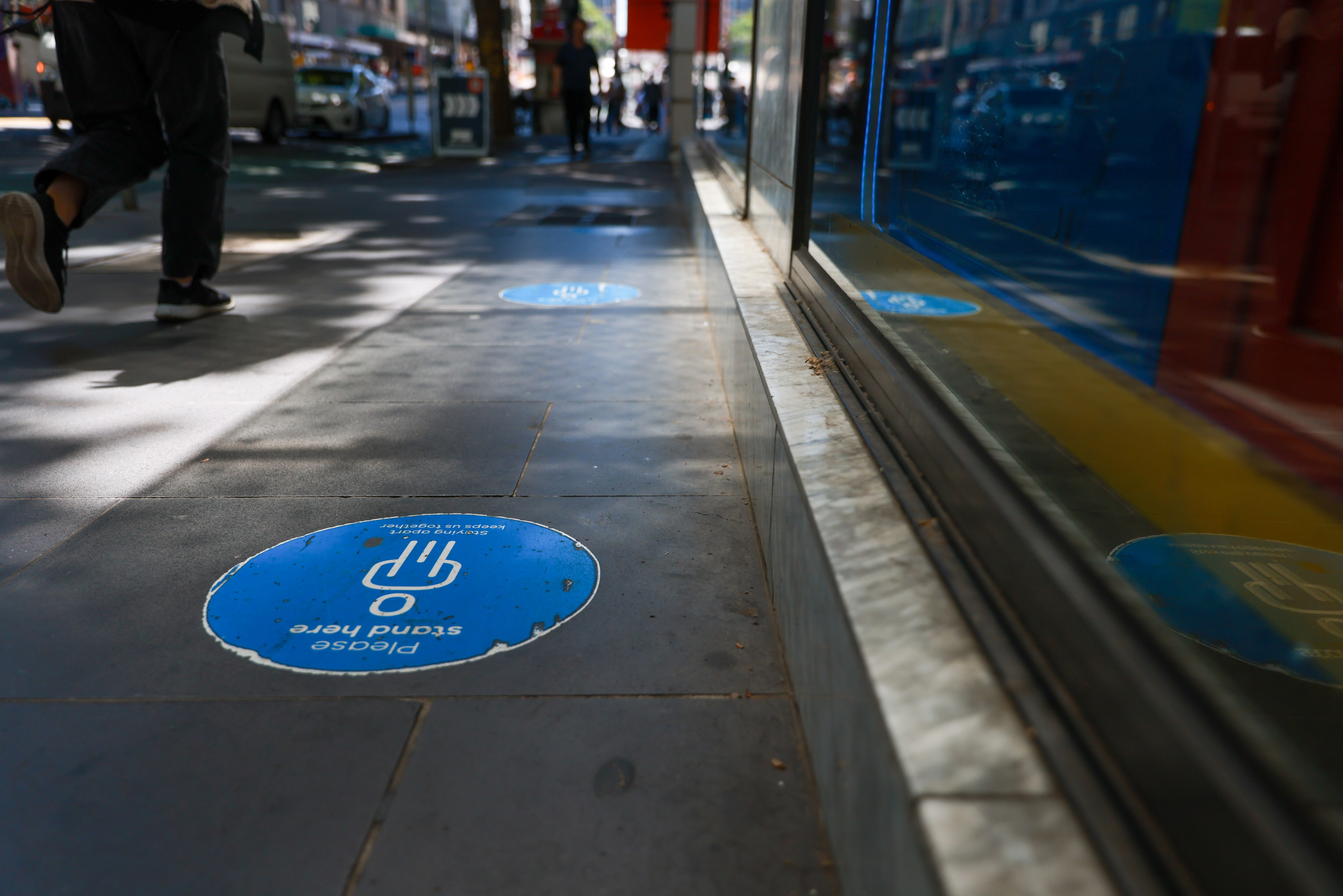 Faded social distance stickers on the footpath and people walking with masks as they pass hand sanitiser stations.