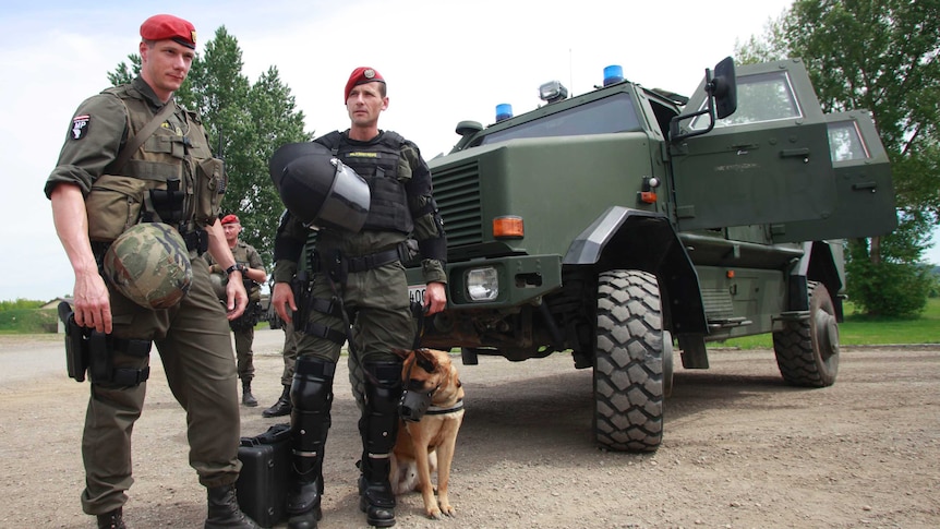 Two men in military gear stand with a dog in front of an army vehicle.