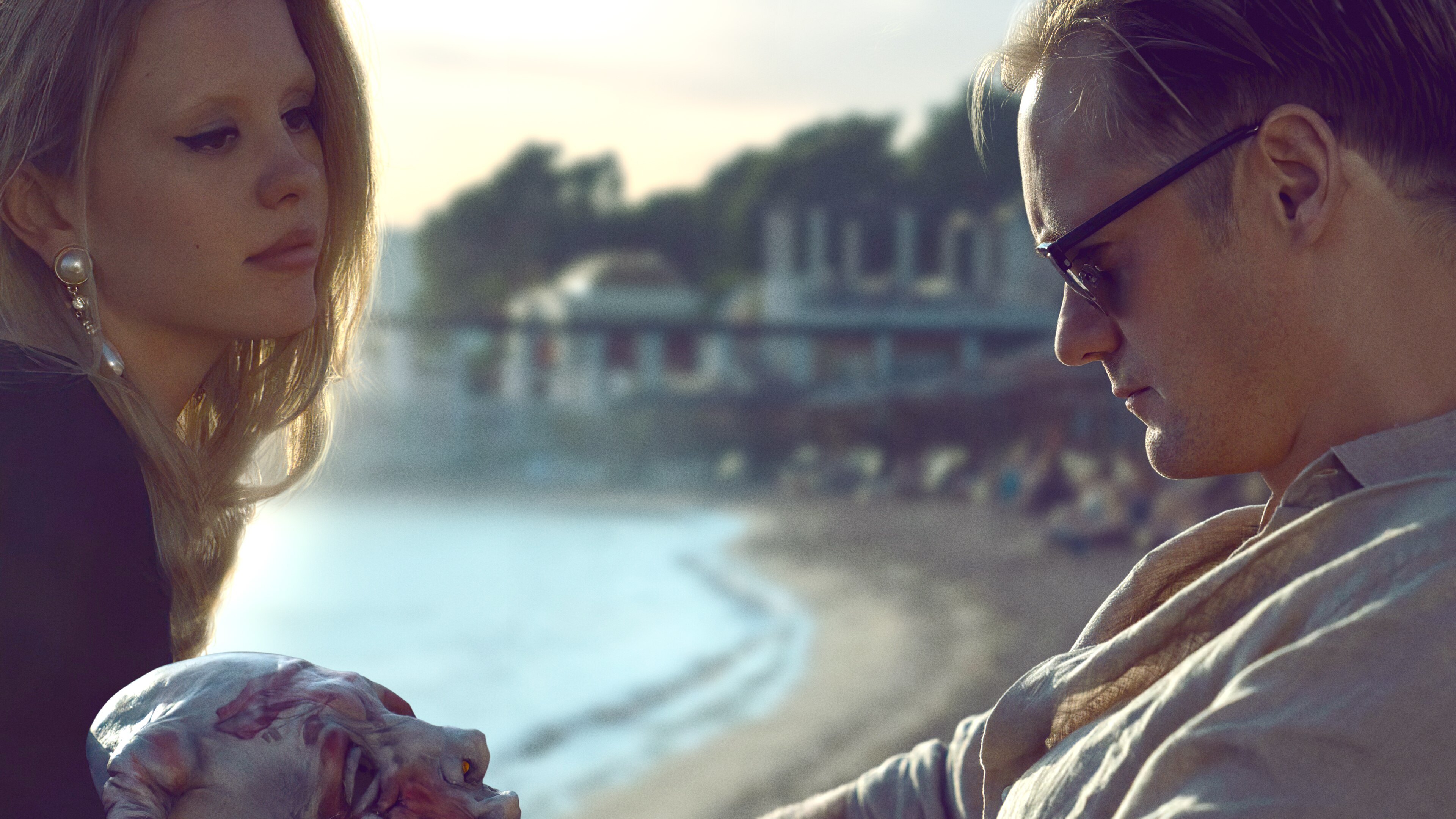 Mia Goth, a blonde white woman with brown eyes, sits on a beach with Alexander Skarsgård, a blonde white man in sunglasses.