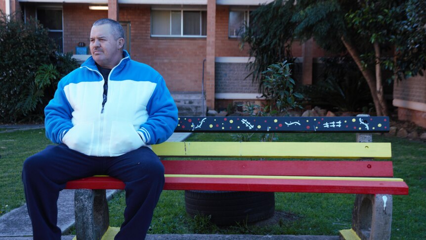 A man wearing a white and blue sweat shirt sits on a park bench