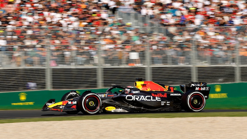 A medium side-shot of a Formula 1 race car driving on a track. A large fence and crowd in stands in the background.