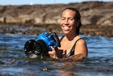 A smiling young woman in the ocean, holding blue underwater housing for her camera.