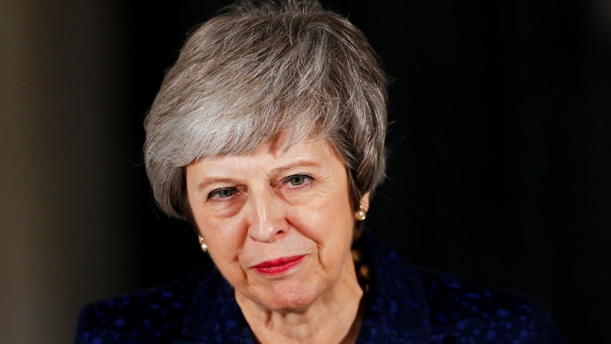 Theresa May stands with pursed lips, looking directly at the camera