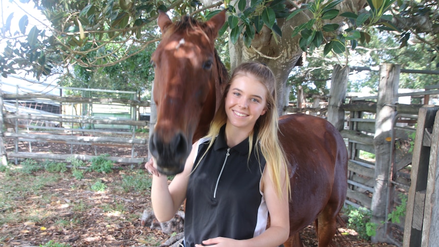 A brown horse standing behind Libby Wylie in a horse yard