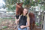 A brown horse standing behind Libby Wylie in a horse yard