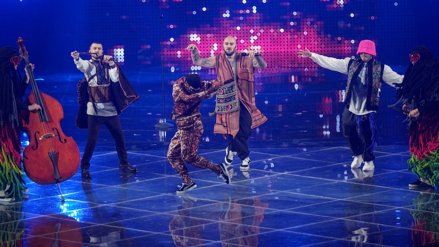 A group of Ukrainian performers in traditional clothing play instruments and dance as one of them break dances in front.