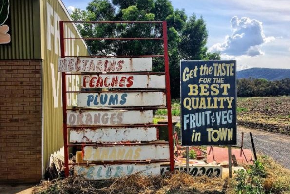 Old faded fruit and vegetable signs on the side of a country road with dry clumps of grass around the bottom of the signs