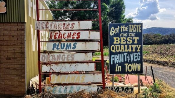 Old faded fruit and vegetable signs on the side of a country road with dry clumps of grass around the bottom of the signs