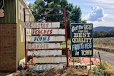 Old faded fruit and vegetable signs on the side of a country road with dry clumps of grass around the bottom of the signs