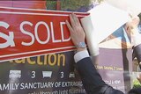 Photo shows hands peeling a sold sticker and placing it on a house for sale sign.