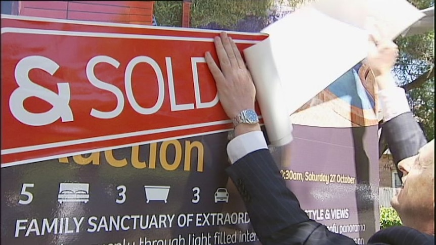 Photo shows hands peeling a sold sticker and placing it on a house for sale sign.