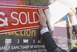 Photo shows hands peeling a sold sticker and placing it on a house for sale sign.