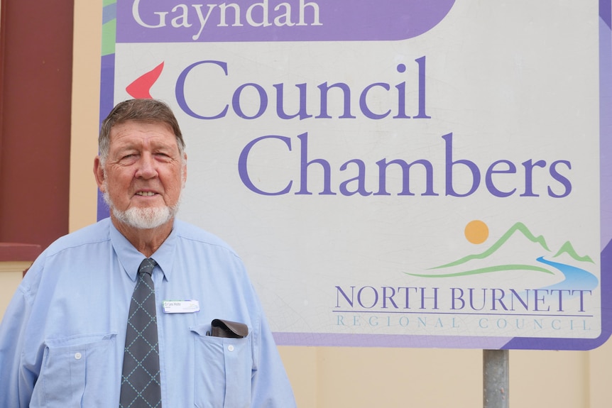 An older man smiles at the camera with a sign behind him reading "Gayndah Council Chambers"