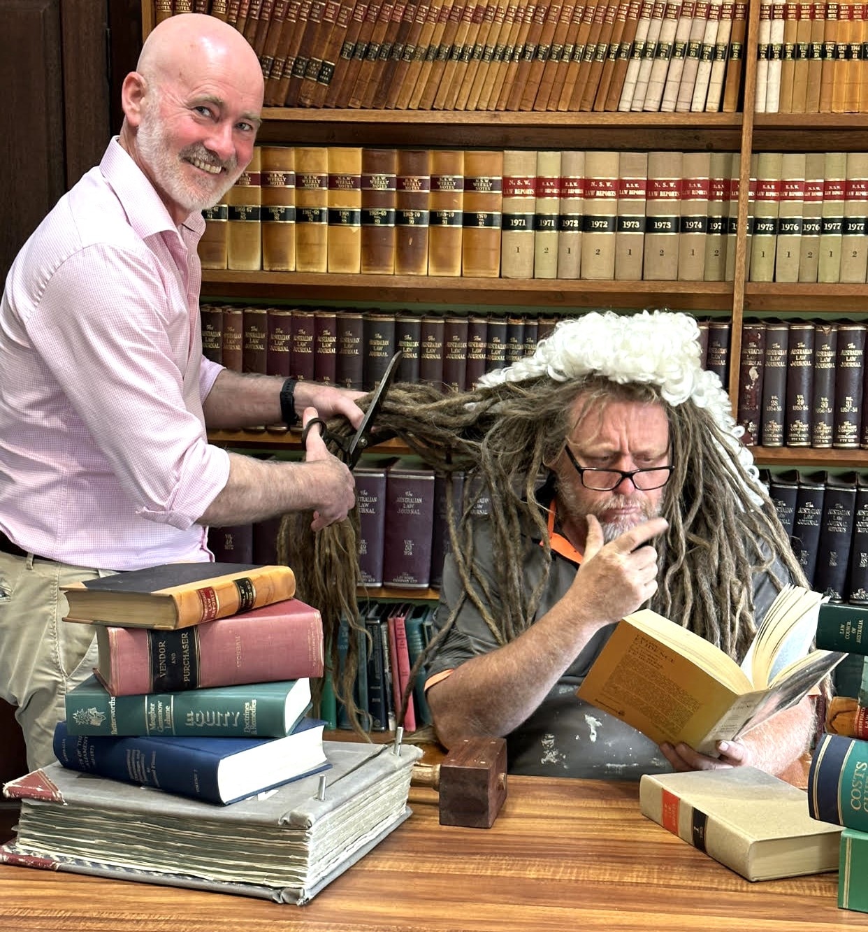 Jason poses as a judge, reading a book, as a lawyer chops his hair