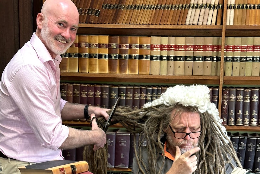 Jason poses as a judge, reading a book, as a lawyer chops his hair