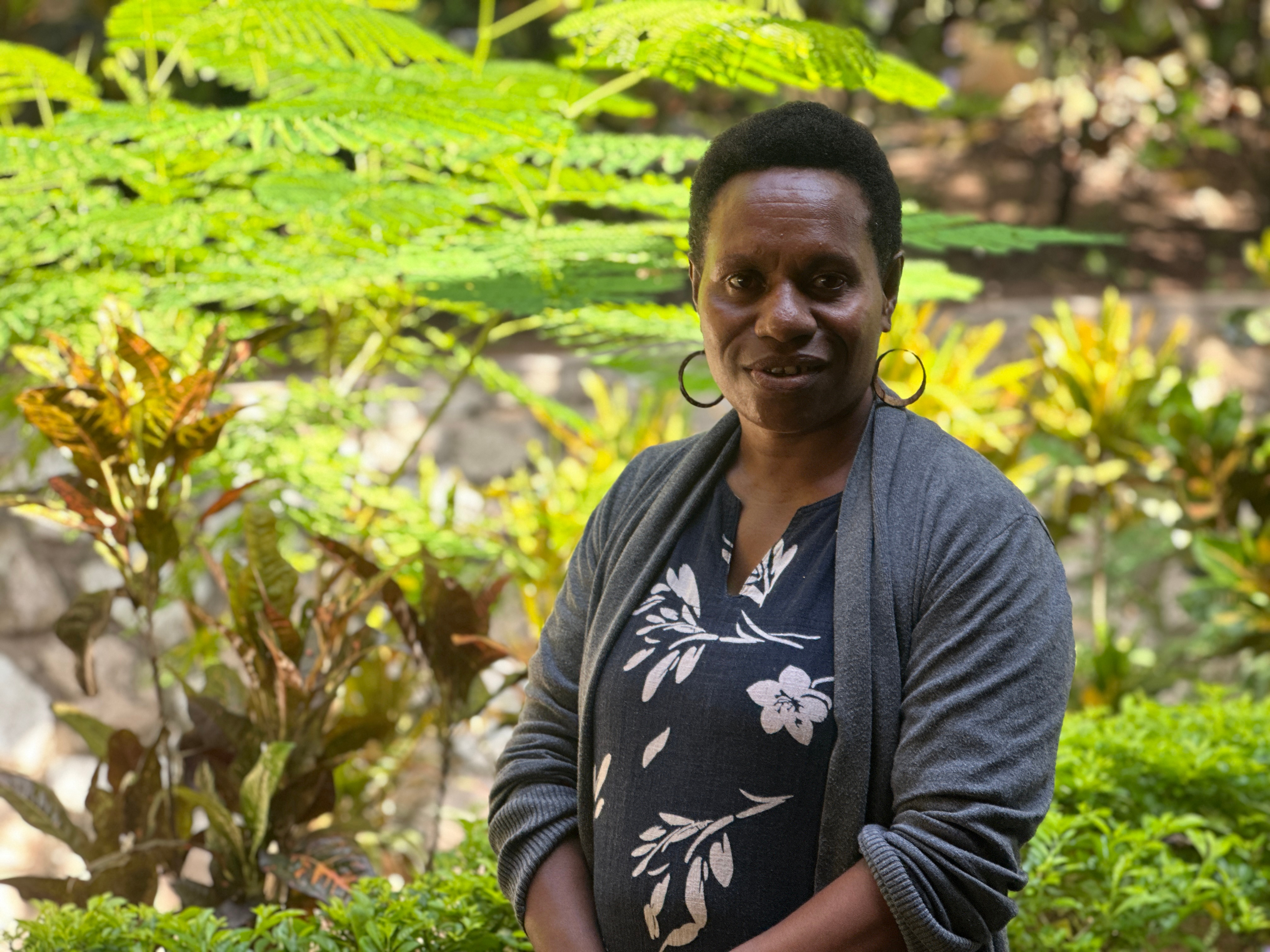 Una mujer sonríe y se para frente a palmeras verdes. Lleva una camisa gris y negra.