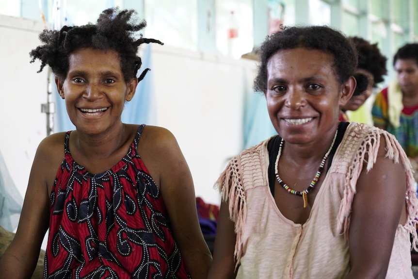 Two women sit next to each other on a hospital bed and smile at the camera.