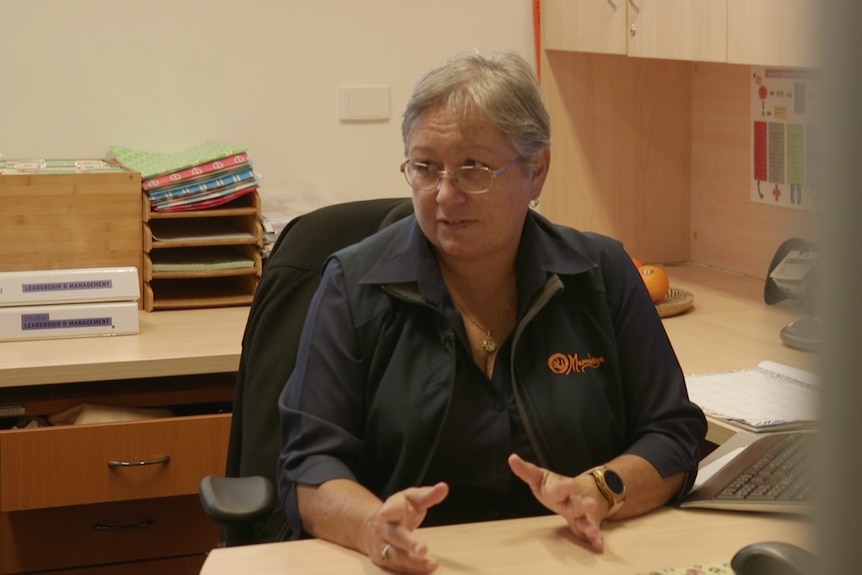 A woman in a black business shirt sits at a desk, chatting to someone out of frame.