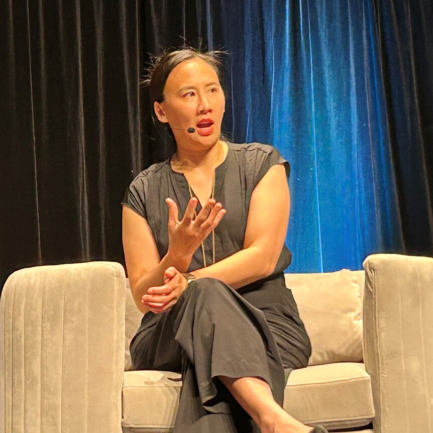 Celeste Ng in black shirt and trousers, seated on taupe couch on stage