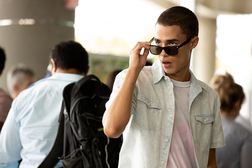 A man wearing light denim short-sleeve shirt over t-shirt, slides sunglasses down nose to inspect something in the distance.